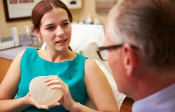 A woman in a teal dress holds a breast implant while discussing with a doctor in a medical office.