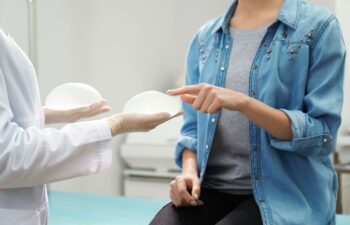 A doctor wearing gloves shows two breast implants to a seated woman in a denim shirt, who is pointing at one of the implants during a consultation.