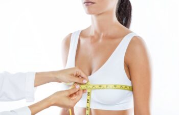 A woman in a white bra is having her bust measured with a yellow measuring tape by another person in a white coat.