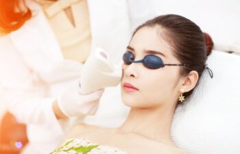 A woman wearing protective goggles receives a facial treatment with a handheld laser device from a professional in a white coat.