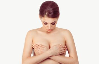 A topless woman covers her chest with her hands while looking downward against a plain white background.