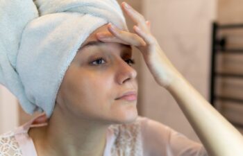 Woman with a towel wrapped around her head touches her forehead while looking in a mirror, wearing a light robe in a bathroom setting.