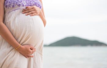 A pregnant person in a light dress stands near water, holding their belly with both hands. An island or hill is visible in the background.