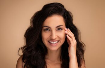 Woman with long dark wavy hair smiles at the camera, touching the side of her face with one hand, against a plain beige background.